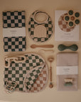 Soft, neutral-toned baby items, including mushie’s Organic Cotton Muslin Burp Cloth 2-Pack, bibs, utensils, a teether, pacifier clip, silicone toy, and folded fabrics—all with minimalist and checkered designs.