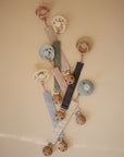 Five FRIGG Rope Silicone Pacifier 2-Packs by FRIGG, each with a pastel-colored clip and wooden clasp in blue, green, black, white, or pink, are arranged on a beige background. All pacifiers are made from medical grade silicone.