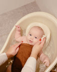 A baby rests in a small tub, covered by a brown towel, while an adult gently washes the baby's head using mushie Muslin Cotton Washcloths. The baby gazes upward, calm and content.