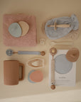 A flat lay of neutral and pastel baby items, including mushie Cradle Cap Brushes with soft silicone bristles—perfect for bath time—plus pacifiers, teethers, a sippy cup, bibs, washcloths, a pacifier clip, spoon, and packaged muslin crib sheet.