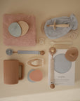 A neatly arranged flat lay of baby essentials, including bibs, pacifiers, a sippy cup, a pacifier clip, washcloths, a teether, Mushie Organic Cotton Bath Mitt for sensitive skin, and a crib sheet—all in soft neutrals and pastels on beige.