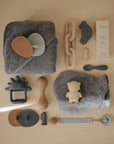A neatly arranged flat lay of baby care items—including the mushie Finger Toothbrush, gray towel, wooden and silicone brushes, toy car, links, teether, pacifier, lotion box, utensils, bib, and plush mitten—on a beige background.