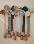 A row of mushie Linen Pacifier Clip 2-Packs, featuring soft pastel linen and smooth wooden clasps in decorative shapes and muted tones, are neatly arranged against a light background.
