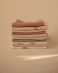 A neatly folded stack of mushie Muslin Cotton Washcloths in soft, neutral tones rests on a beige surface with a minimalist background. Some cloths feature subtle patterns or small embroidered details.
