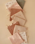 The mushie Muslin Cotton Washcloths, in soft neutral, pink, and beige tones with simple patterns like rainbows or flowers and solid colors, feature a fabric tag and loop. They are small, square, and arranged diagonally.