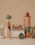 Aesthetic arrangement of mushie Muslin Cotton Washcloths with pastel baby care essentials—bottles, stackable toys, towels, bibs, brush, pacifier, spoons, and cups—displayed on a neutral beige background.