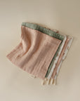 Three muslin cotton washcloths by mushie—organic, pastel pink, soft green, and white floral—are neatly folded on a neutral background, with cream-colored tags showing on one edge.