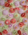 Mushie’s FRIGG Rope Natural Rubber Pacifier 2-Pack in pink, red, and green lies scattered on a pale yellow surface adorned with daisies and floral illustrations.