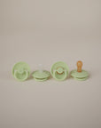Four FRIGG Rope Natural Rubber Pacifiers by Mushie, a baby essential, are displayed in a row. Each has a unique natural rubber latex nipple style or color—green, clear, yellow, and orange—included in two-packs.