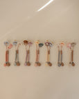 Four pastel-colored FRIGG Natural Rubber Pacifier 4-Packs, each attached to a beaded clip with a smiling animal face, are arranged in a row on a light surface.