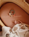 A woven baby bassinet with a soft pink sheet, a silicone teething ring toy, a pink pacifier with a beaded clip, and a light, striped blanket draped inside.