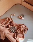 A soft beige blanket, a gray toy ball, a beaded pacifier clip, and a teething ring rest on a light gray crib mattress with a wooden frame.