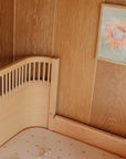 A wooden crib fitted with a mushie Stretchy Crib Sheet, patterned with small hearts, stands against wood-paneled walls. Above the crib, a framed picture of a smiling sunflower decorates the wall.