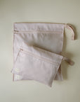 Two light pink mushie Water Resistant Wet Bags with wrist straps lie on a cream surface. The larger bag overlaps the smaller; both have a subtle checkered design, ideal for diaper bags or organizing essentials.