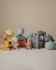 A pastel-themed display features mushie's Daisy Rattle Teether alongside whimsical rubber toys shaped like flowers, animals, vehicles, and rockets, all set against a plain beige background.