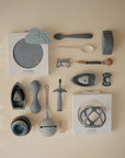 A flat lay of mushie baby feeding and teething essentials in muted blue and gray, featuring food-grade silicone plates, spoons, teething rings, stacking cups, toys, and the mushie Silicone Pacifier Clip | Eva—neatly arranged on a light beige surface.