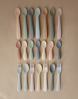 A top-down view of three rows of mushie Silicone Feeding Spoons, arranged in matching pastel color groups on a beige surface.