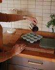 Someone pours liquid from a metal measuring cup into the Mushie Baby Food Freezer Tray, made from BPA-free silicone, on a wooden kitchen countertop with tiled walls and a small plant—ideal for convenient baby food storage.