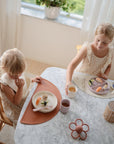 Two young children wear mushie Silicone Baby Bibs while eating at a marble table by a window.