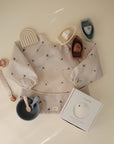 Flat lay of baby items on beige, featuring a mushie Long Sleeve Bib (water-resistant), silicone bowl with spoon and fork, pacifier clip, toy boats, rainbow toy, and boxed mushie bib.