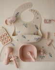 A flat lay of pink and cream baby feeding accessories by mushie, including the Silicone Pacifier Clip | Hera, a bib with blue flowers, bowl, spoon, teethers, teething ring, and pop toy—all arranged on a matching floral mat.