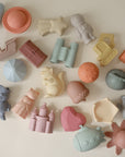 The mushie Nature Mold Free Bath Play Set features colorful, food-grade silicone toys—animals, castles, a heart, binoculars, a boat, and an astronaut—in pastel blue, pink, orange, green, and yellow scattered on a light surface.