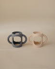 The mushie Silicone Bottle Handles—one dark gray, one light beige—feature wide grips and work as universal baby bottle accessories. They sit side by side on a softly lit, neutral background.