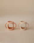 Two mushie Silicone Bottle Handles are shown side by side on a neutral background—one in soft peach, the other in light beige. Both feature large handles and a minimalist, modern design perfect for use as baby bottle accessories.