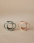 Two mushie Silicone Bottle Handles, each with three handles and in muted green and pale pink, are displayed side by side on a beige background—ideal as universal bottle or baby bottle accessories.