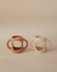 Two mushie Silicone Bottle Handles, one light brown and one beige, are side by side on a neutral beige background. These universal baby bottle accessories have open sides and a minimalist design.