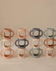 Six pastel Silicone Bottle Handles by mushie, with cut-out centers and double handles, are stacked in two rows. The beige, pink, blue, and green colors pop against the neutral background.