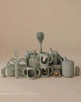 A playful set of pale green kitchenware by mushie is arranged on a beige background, featuring mugs, a spoon, whimsical designs, and Silicone Bottle Handles for sippy cups.