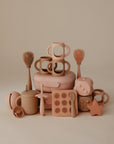 A tidy display of mushie silicone baby toys and accessories in gentle hues, featuring Silicone Bottle Handles, cups, brushes, a divided tray, dice, a star, a pacifier, and other playful shapes.