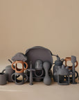 A selection of mushie’s matte, pastel silicone baby items—cups, plates, utensils, teethers, toys, and Silicone Bottle Handles—arranged neatly on a beige background. Some pieces have fun shapes like cars and clouds.