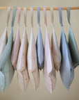 A row of mushie Water Resistant Wet Bags in pastel shades—light blue, pink, cream, and mint green—hang by straps from a wooden rod. Ideal for diaper bag storage and keeping essentials organized in style.