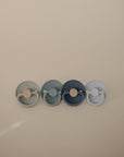The FRIGG Silicone Pacifier 4-Pack features four round pacifiers in muted green, blue, and gray shades, arranged on a beige background. Made by FRIGG from medical grade silicone for your baby’s comfort.