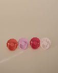 The FRIGG Silicone Pacifier 4-Pack by FRIGG features four medical-grade silicone pacifiers in red, pink, purple, and white. Each has a round handle and a scalloped shield, shown lined up on a neutral beige background.