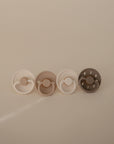 Four FRIGG Natural Rubber Pacifiers by FRIGG, shown in white, tan, cream, and dark olive on a beige background. Each pacifier is crafted in Denmark with natural rubber and features unique braided rope and moon phase handle designs.