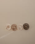 The FRIGG Silicone Pacifier 4-Pack by FRIGG features four minimalistic pacifiers in white, beige, and brown tones with braided and crescent moon details, crafted from medical-grade silicone. They’re shown neatly arranged on a soft beige background.