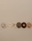 Six FRIGG Silicone Pacifiers in assorted neutral tones are neatly lined up side-by-side on a plain light beige background.