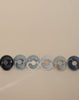 The FRIGG Natural Rubber 6-Pack by FRIGG includes six pacifiers in blue and gray with plain, rope, and moon and star designs, all made in Denmark and displayed against a beige background.