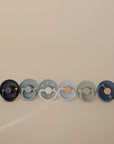 Six FRIGG Silicone Pacifiers in a 6-pack set, featuring assorted blue and gray shades, are shown arranged on a beige background. Made in Denmark, each pacifier has a round shield with unique textures and details.