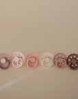 The FRIGG Silicone Pacifier 6-Pack by FRIGG features six medical grade silicone pacifiers in pastel purple, pink, beige, and brown. Each has a scalloped edge and is beautifully arranged against a neutral background.
