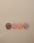 The FRIGG Natural Rubber Pacifier 4-Pack by FRIGG features four pastel pacifiers with scalloped edges and heart-shaped cutouts, shown in brown, mauve, pink, and lavender against a beige background.