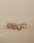 The FRIGG Silicone Pacifier 6-Pack by FRIGG features six medical-grade silicone pacifiers in neutral beige, taupe, and brown tones, each with scalloped or round edges for a minimalist, modern style against a soft beige background.