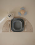 The mushie Dinnerware Cutlery Set includes a non-toxic, dishwasher-safe cup, plastic cutlery, two gray nesting bowls, and a sun-shaped toy with blue clouds on a speckled semi-circle placemat—perfect for your baby’s place setting.