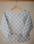 The mushie Long Sleeve Bib, featuring a blue and white checkerboard pattern and water-resistant design, hangs on a wooden hanger against a neutral background.