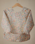 The mushie Long Sleeve Bib features a water-resistant, pastel floral design with a wide pocket. This machine washable bib hangs on a wooden hanger against a neutral background.