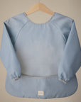 A mushie Long Sleeve Bib in light blue hangs on a wooden hanger against a beige background. This water-resistant baby bib has elastic cuffs, a wipe-clean surface, and a bottom pocket for easy mealtime cleanup.