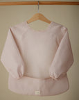 The mushie Long Sleeve Bib in light pink is water resistant, features elastic cuffs, and is displayed on a wooden hanger against a beige background.